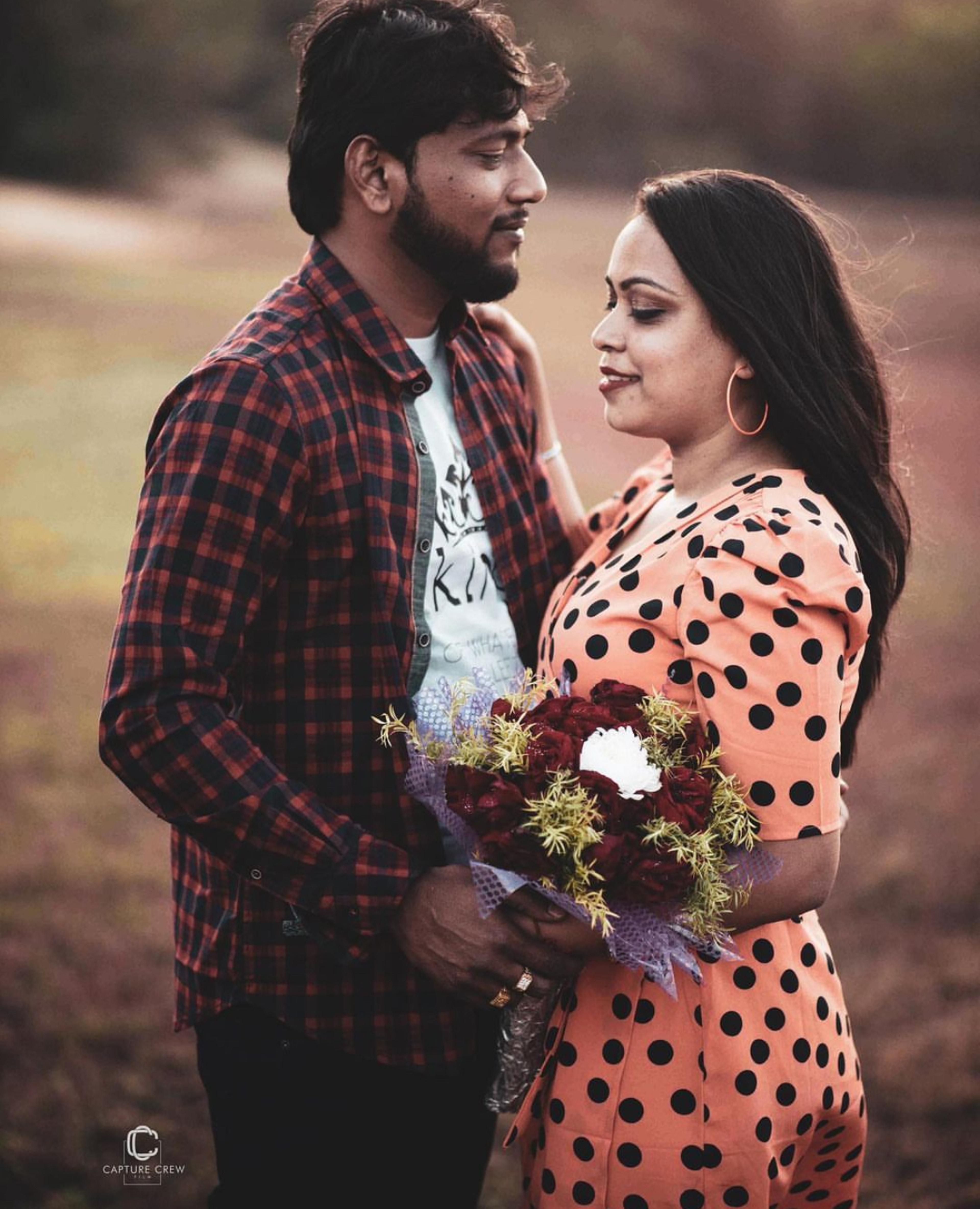 Romantic Couple standing close in a scenic outdoor setting,captured by Capturecrew Films or tccf . The man holds a bouquet of red and white flowers, and the woman in a peach polka-dot dress gazes at him lovingly with a warm , natural background.