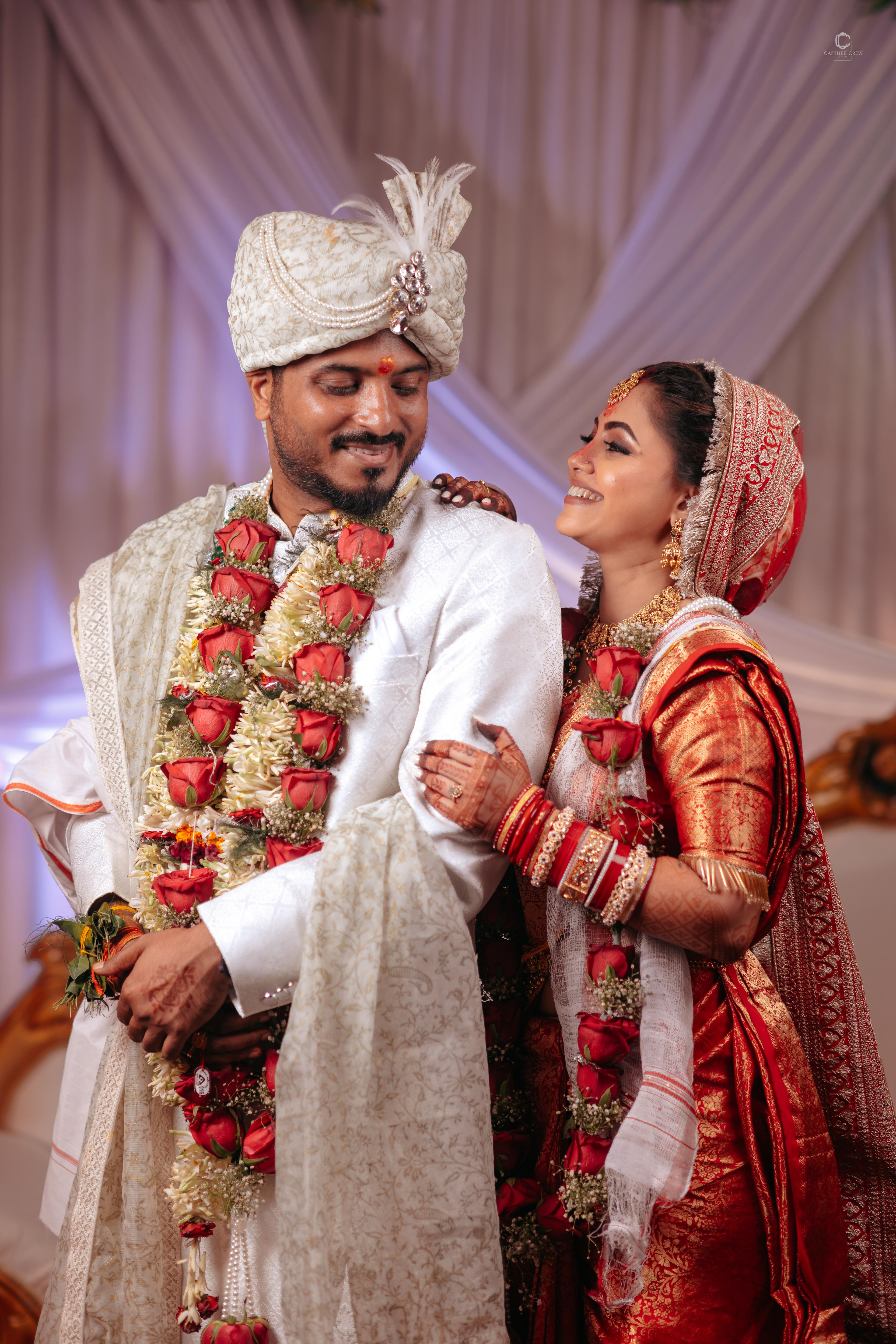 Traditional Indian wedding couple captured by CaptureCrew Films (TCCF). The bride, dressed in a red silk saree with intricate jewelry, and the groom in a white sherwani and turban adorned with floral garlands, share a joyful moment. Expertly captured Indian wedding photography.