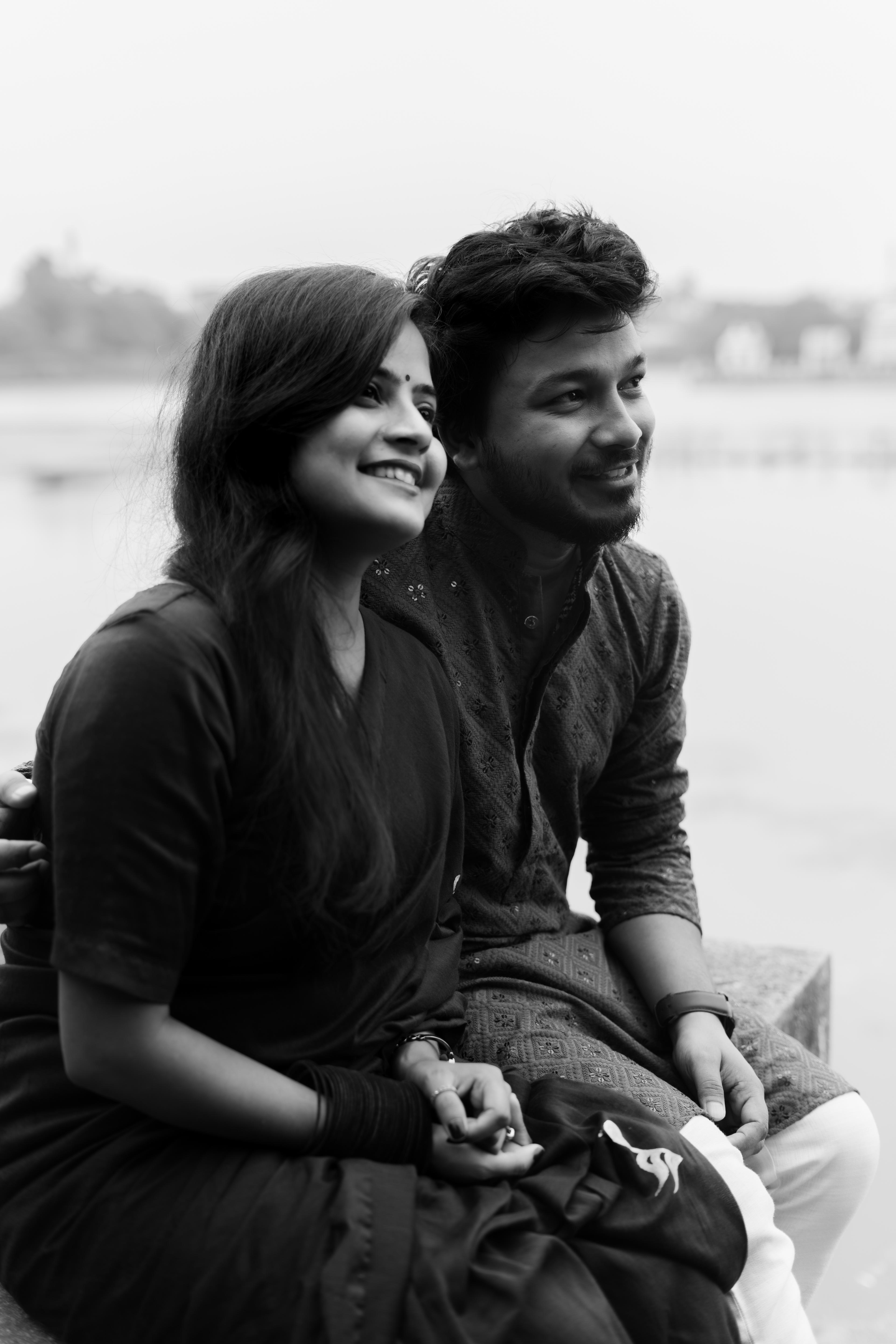 Engaged couple enjoying a candid black-and-white pre-wedding photoshoot by CaptureCrew Films (TCCF). The couple sits close, smiling as they look into the distance, capturing a natural and intimate moment. Expertly crafted pre-wedding photography.